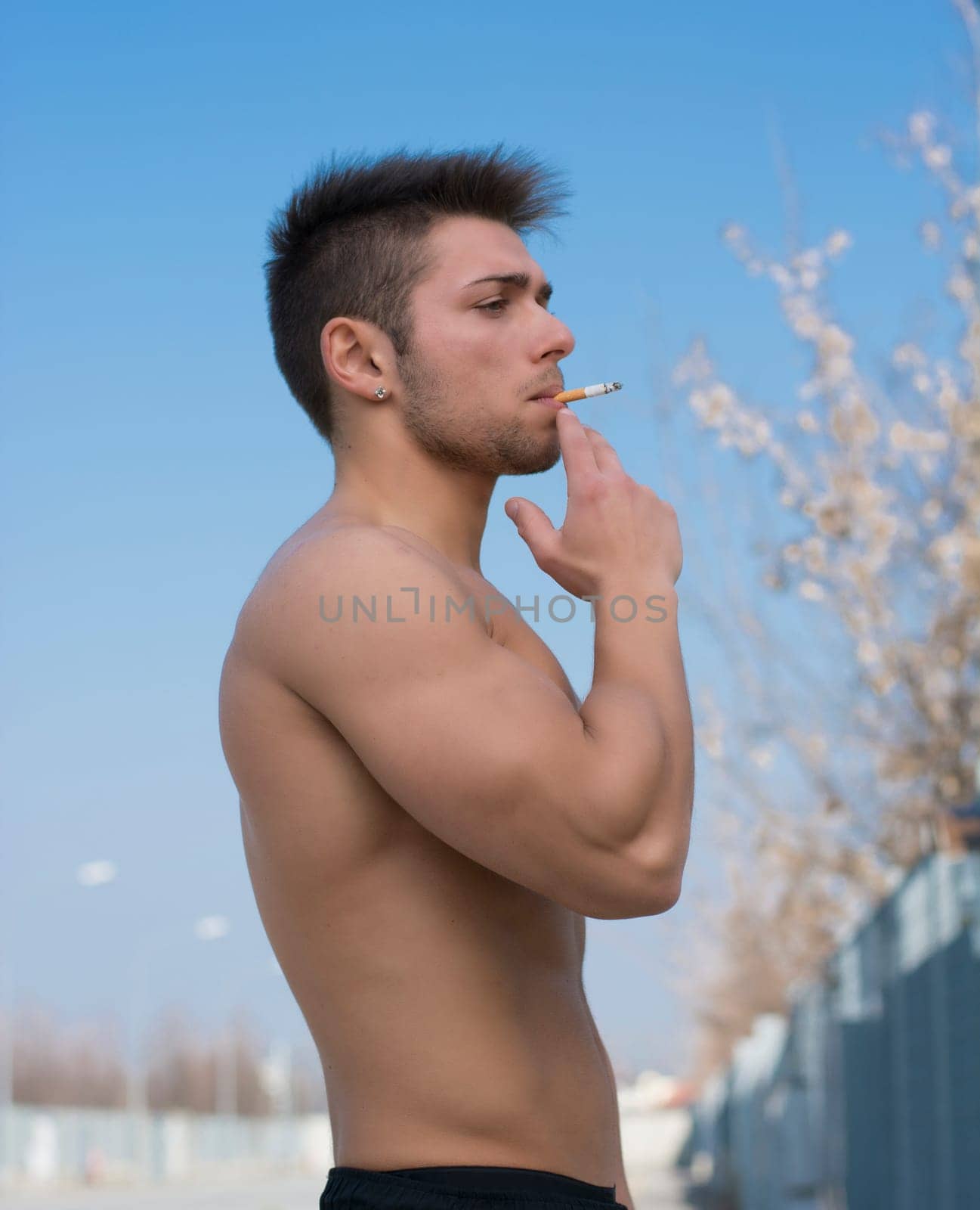 Shirtless muscular young man smoking cigarette outside, standing, profile view