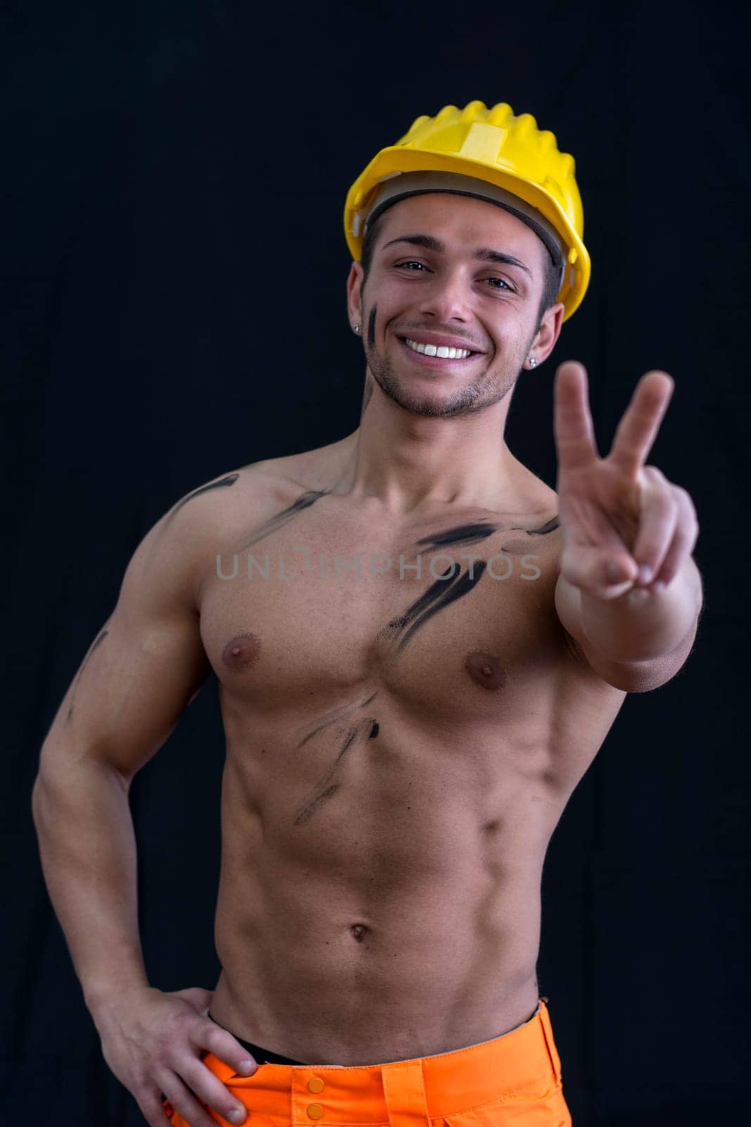 Muscular young construction worker shirtless doing peace sign isolated on black background