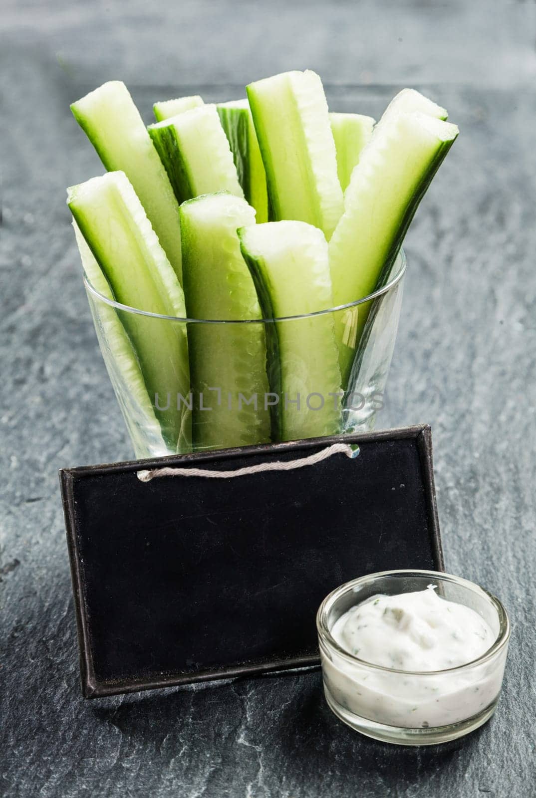 Sliced cucumber batons or crudites served with a small bowl of creamy savory vinaigrette sauce and a blank school slate with copyspace