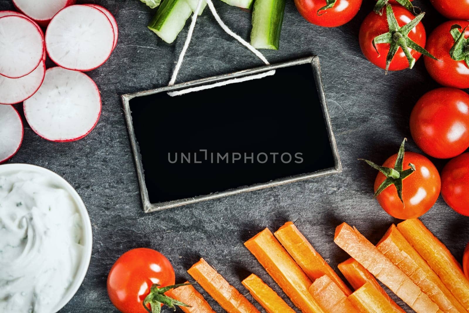 Sliced fresh vegetable frame around a small blank vintage school slate including radish, carrot batons, tomato and cucumber served as crudites with savory dip
