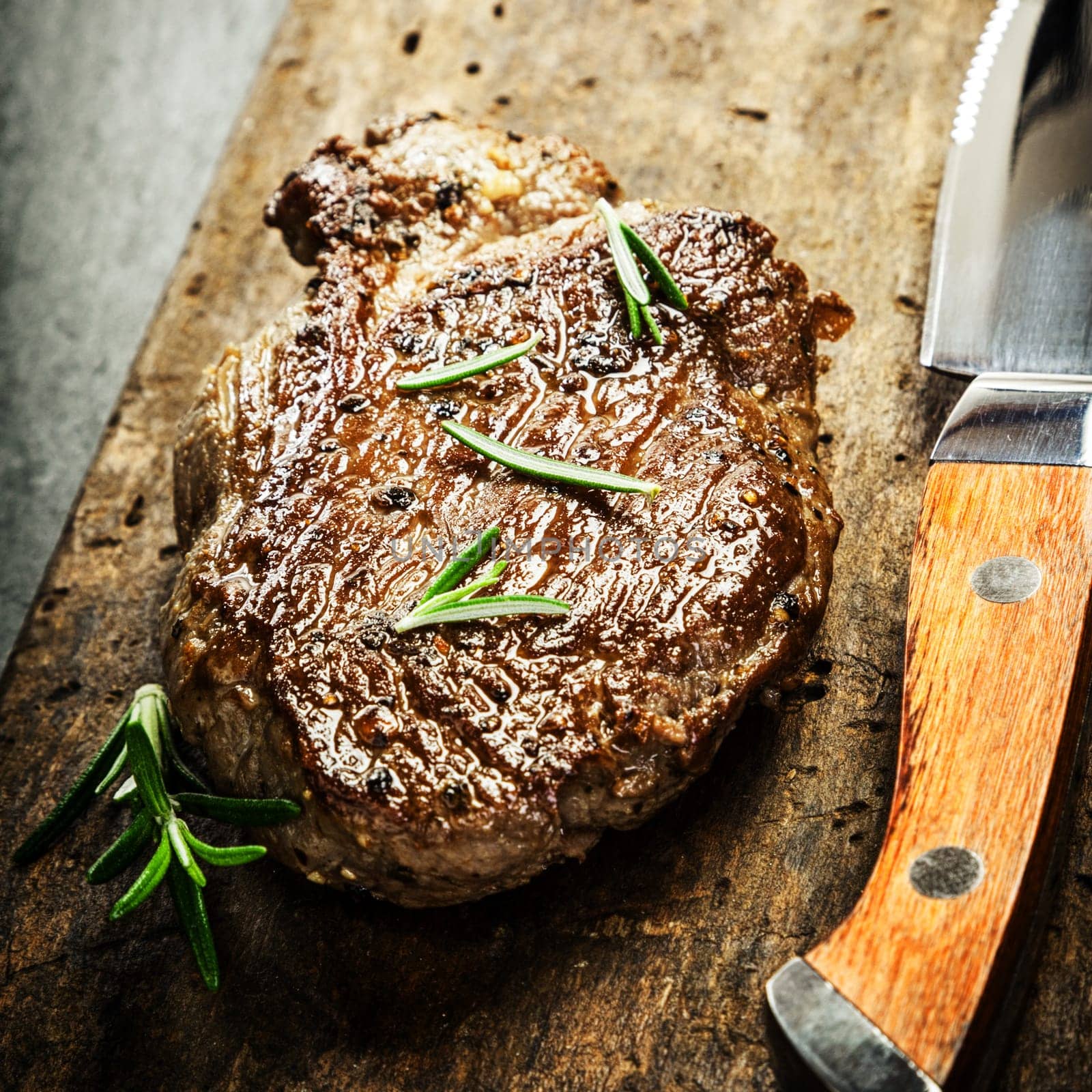 Succulent portion of grilled beef steak garnished with fresh rosemary on a rustic wooden chopping board with a sharp steak knife for a gourmet meal