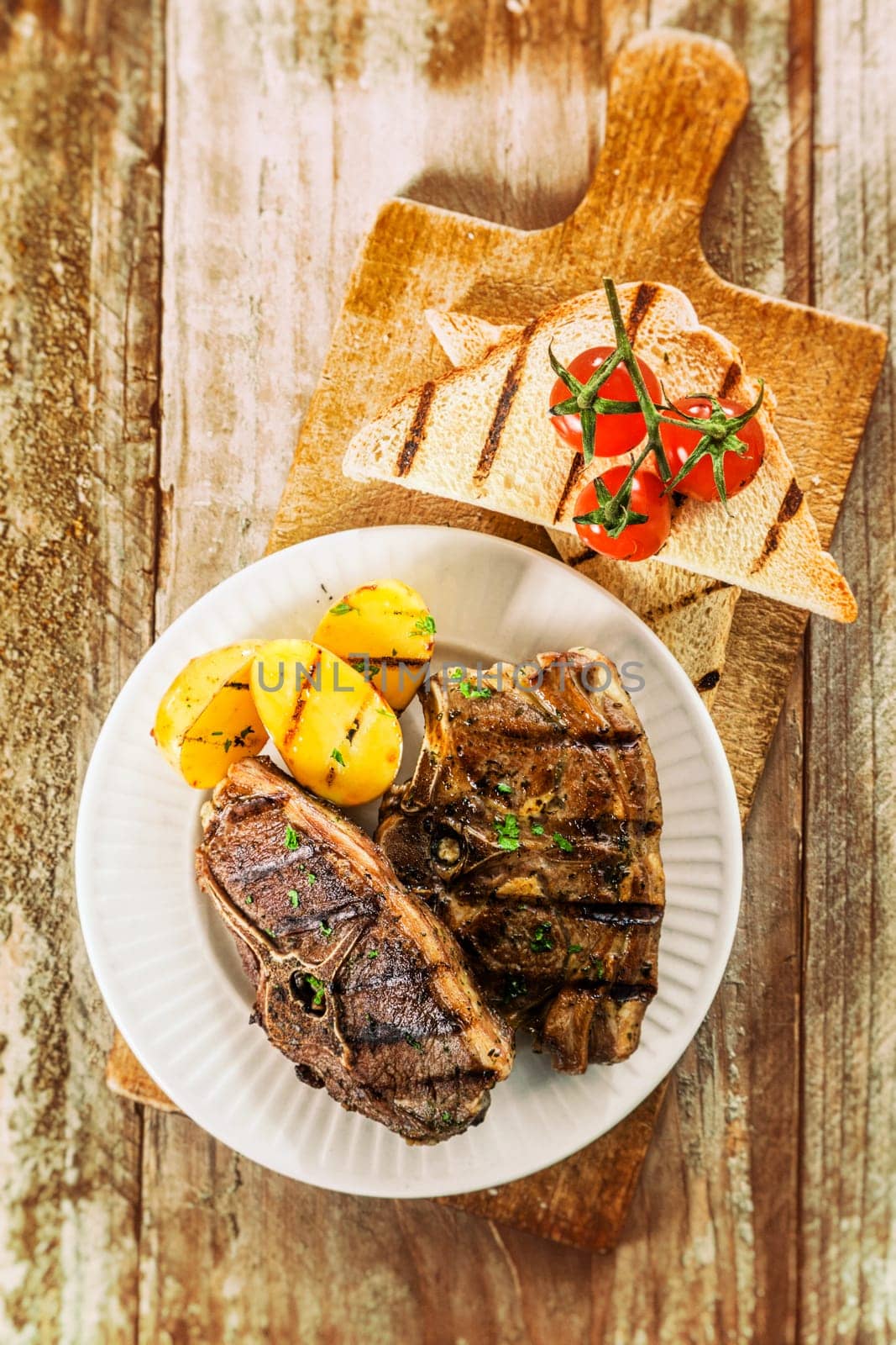 Tender grilled chops off the barbecue served with grilled potato and cherry tomatoes and a slice of toast on an old weathered picnic table, overhead view