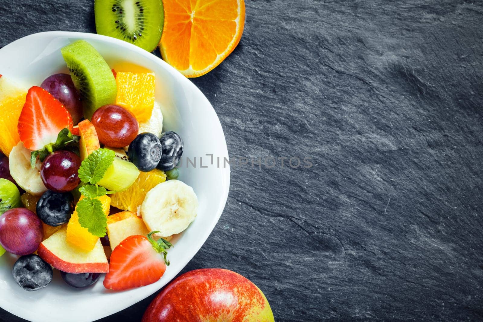 Overhead view of a bowl of fresh fruit salad for a healthy diet made with assorted tropical fruits and an apple, kiwifruit and orange arranged alongside on textured slate with copyspace