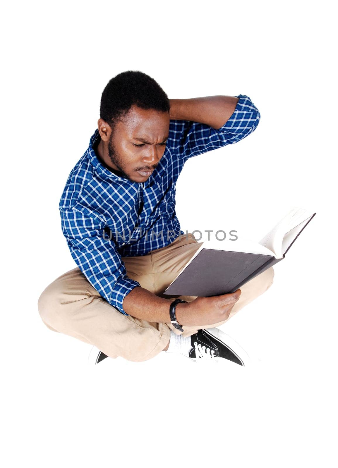 A handsome young African American man in beige pants and a blue.shirt sitting on the floor, reading a book, isolated on white background..