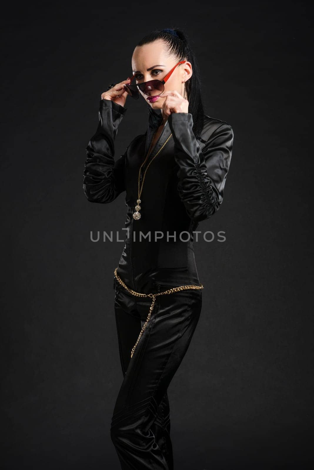 Portrait of beautiful Caucasian woman putting on sunglasses and wearing black bodysuit against black background