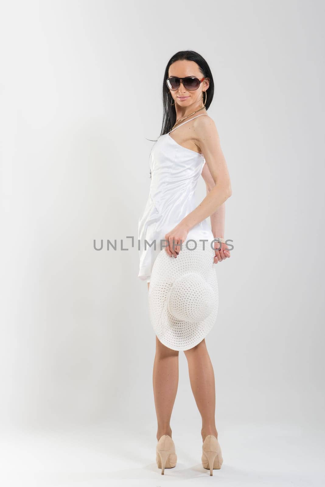 Full length shot of beautiful Caucasian woman wearing short white dress, sunglasses and holding large white hat, turning to look at camera