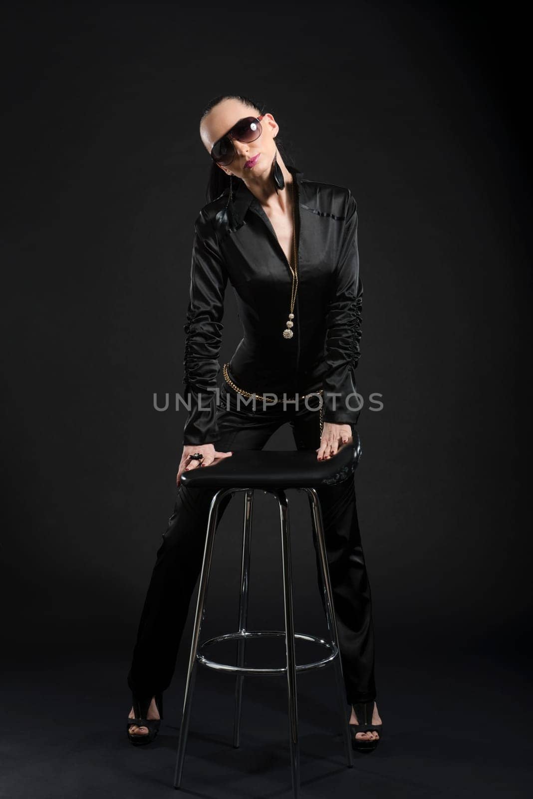 Full length shot of intimidating Caucasian woman wearing black bodysuit and sunglasses, against black background