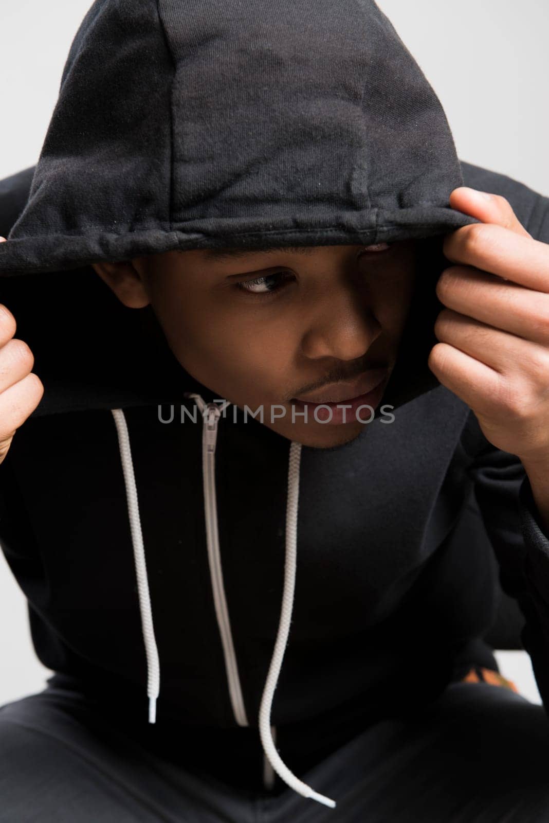 Portrait of suspicious dark skinned male model hiding under black hooded jumper