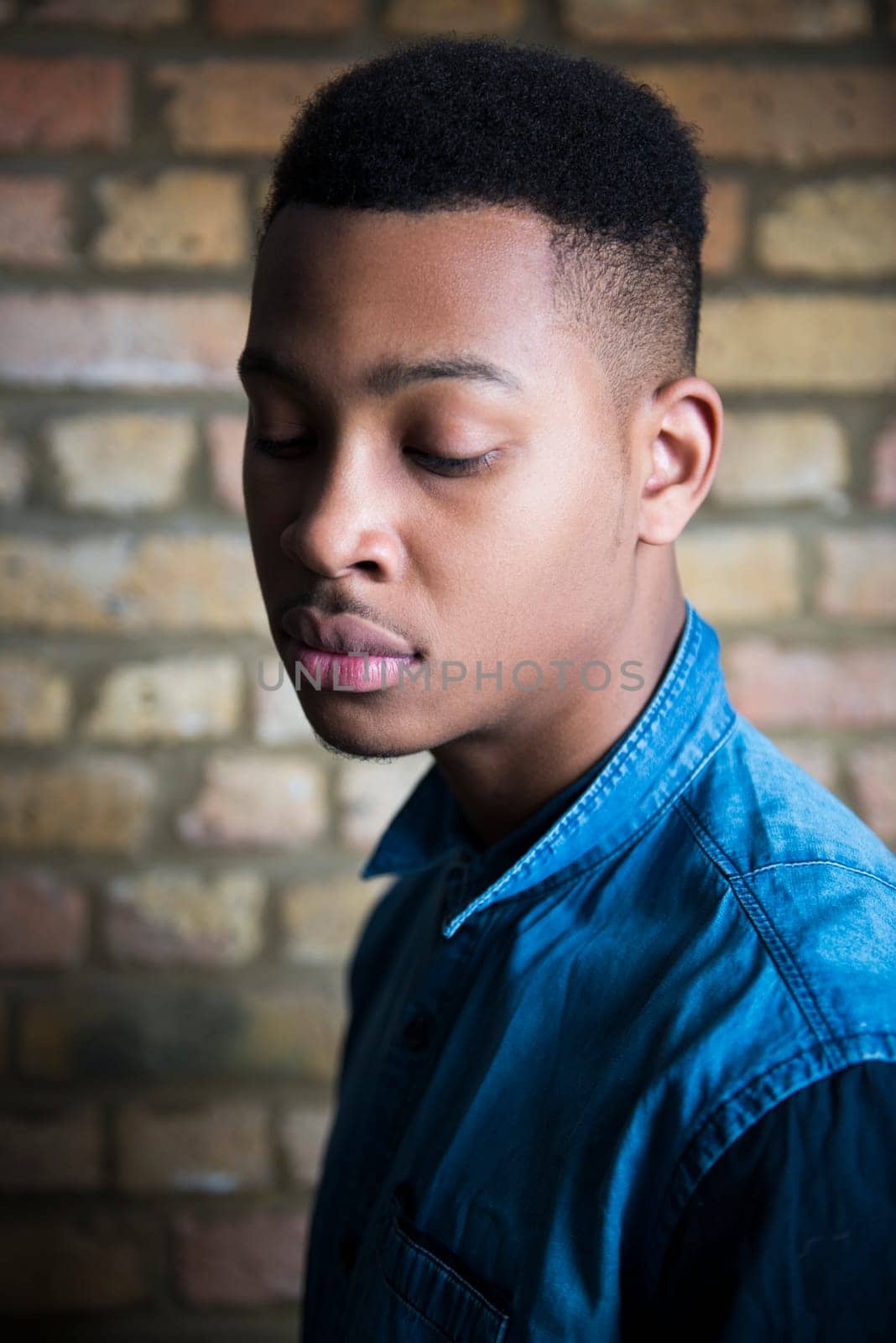 Shallow depth of field portrait of handsome dark skinned male model looking down and wearing casual clothes against brick wall