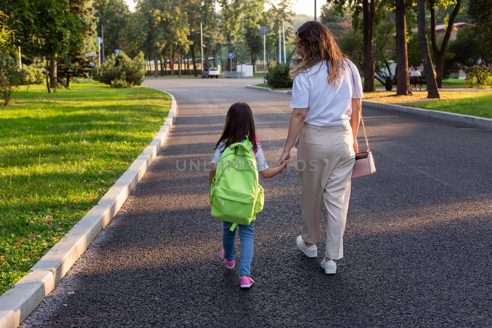 Mother and daughter walking together in park with backpack at sunset.