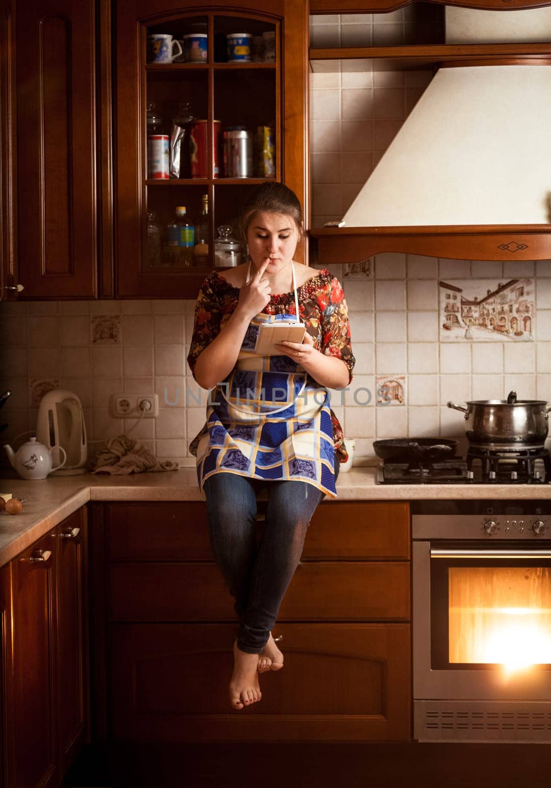 Young housewife sitting on tabletop and looking for recipe in tablet