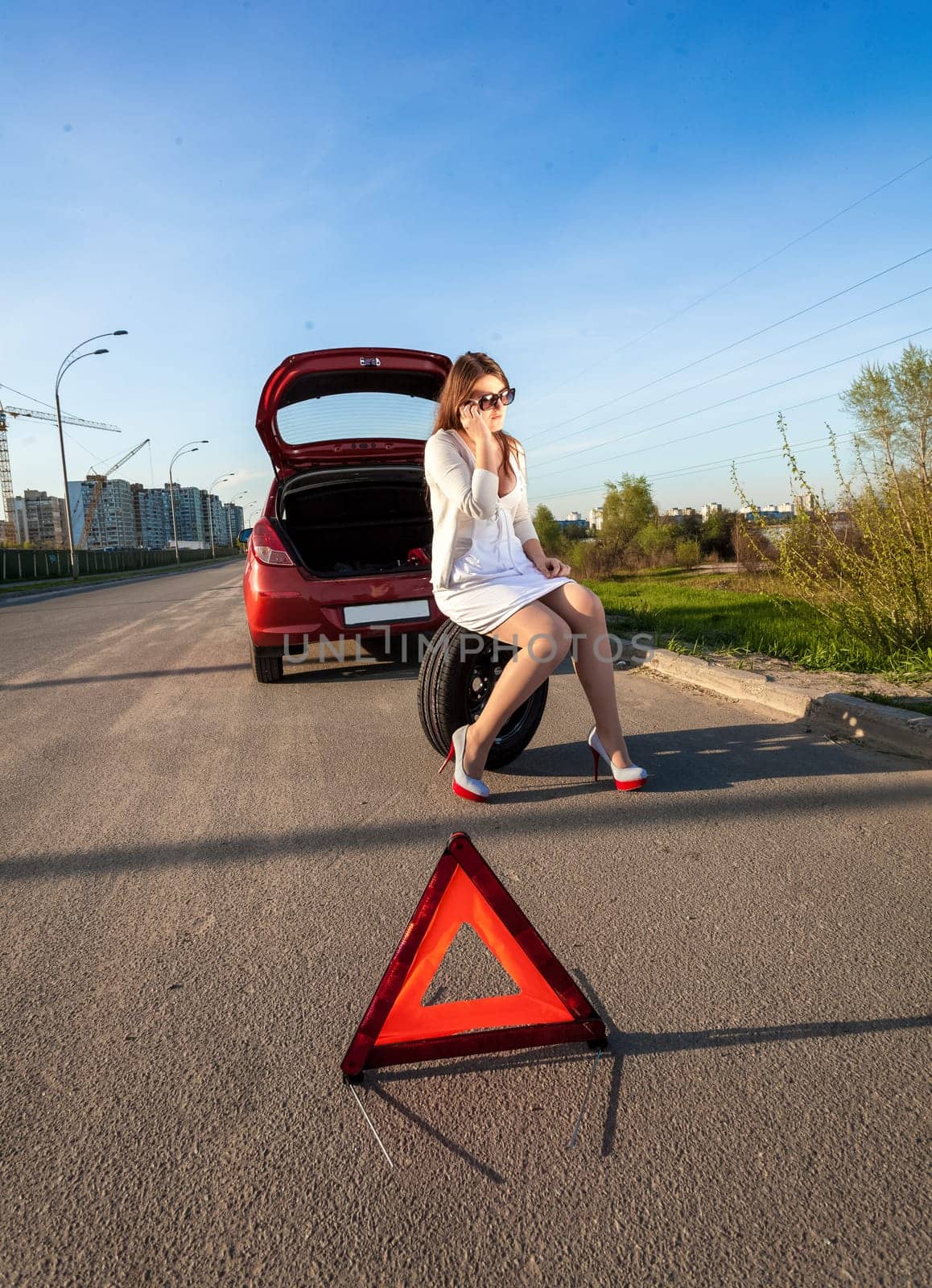 Sexy woman sitting on spare wheel against of broken car and warning sign