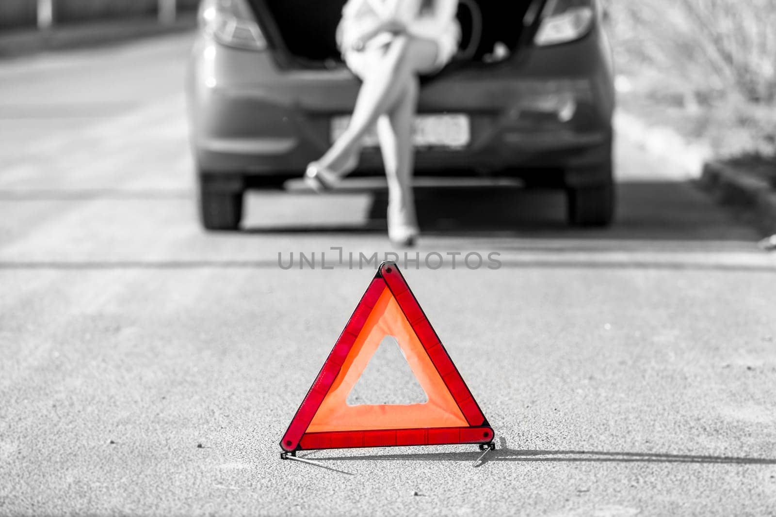 Black and white photo of woman sitting on broken car near red warning sign