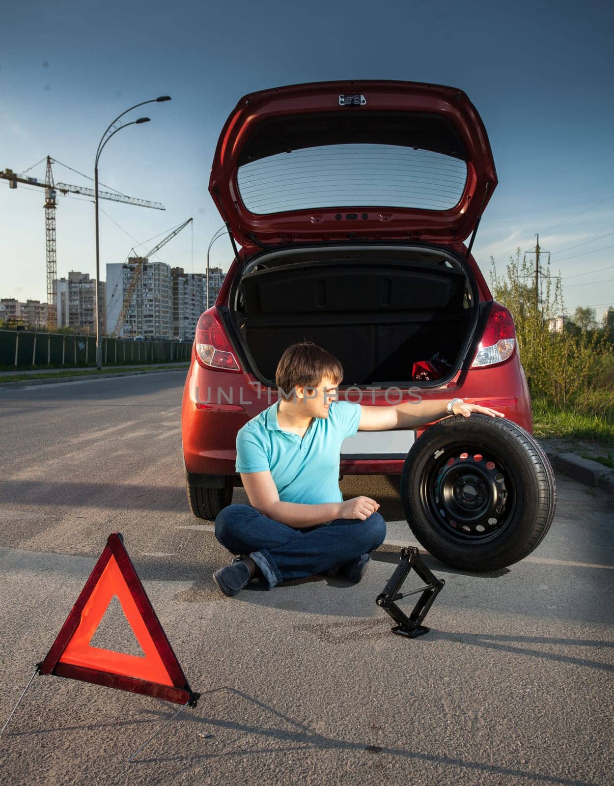 Young man sitting on road and leaning against broken car