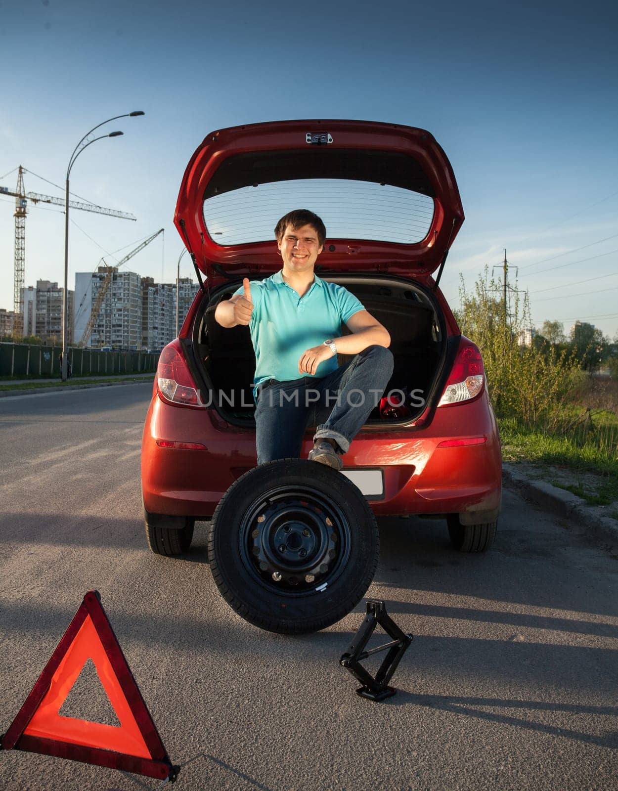Young man sitting on car trunk and holding foot on spare wheel