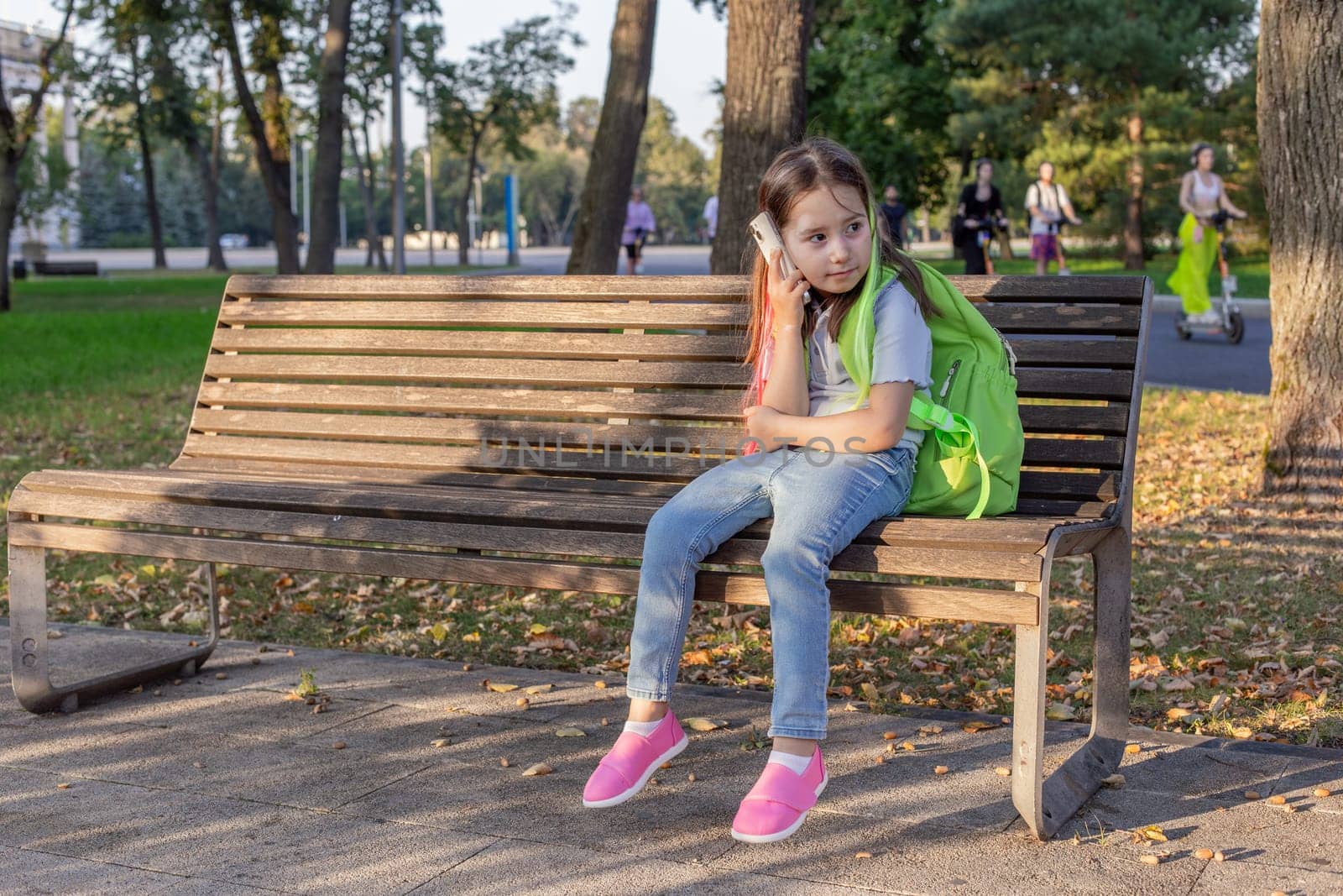 Young girl with backpack sitting on park bench in autumn sunlight.