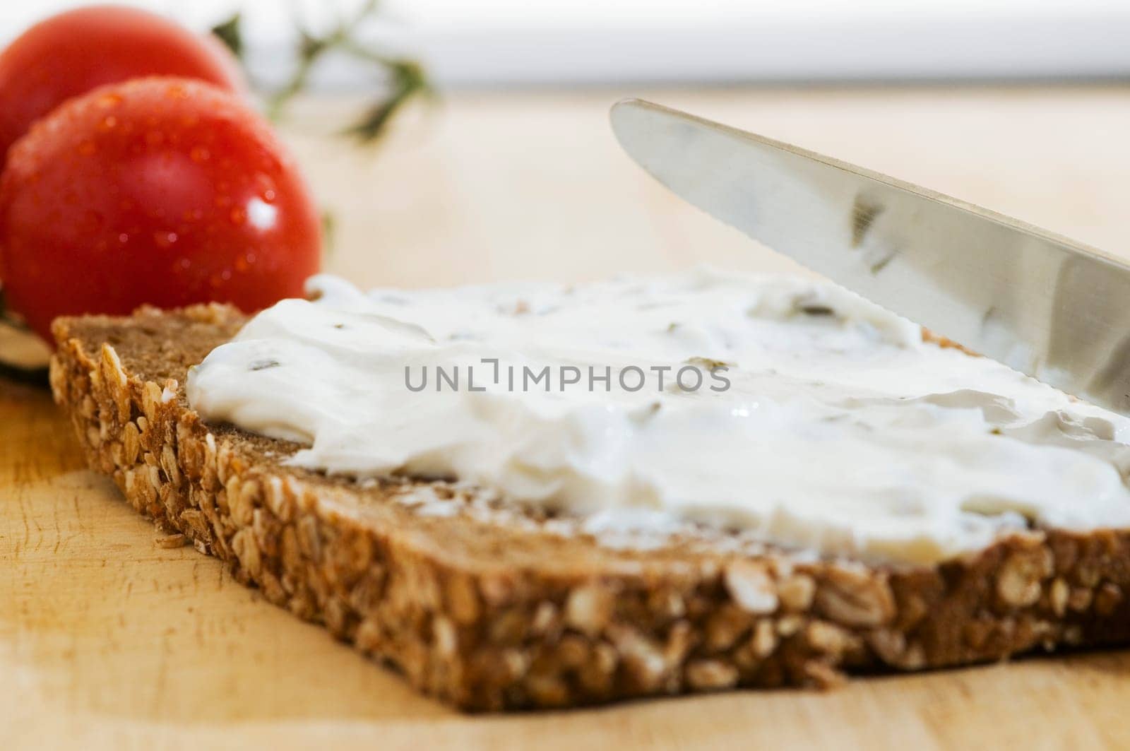 Close-up of a slice of bread covered with white cheese and two tomatoes behind it on a wooden cutting board