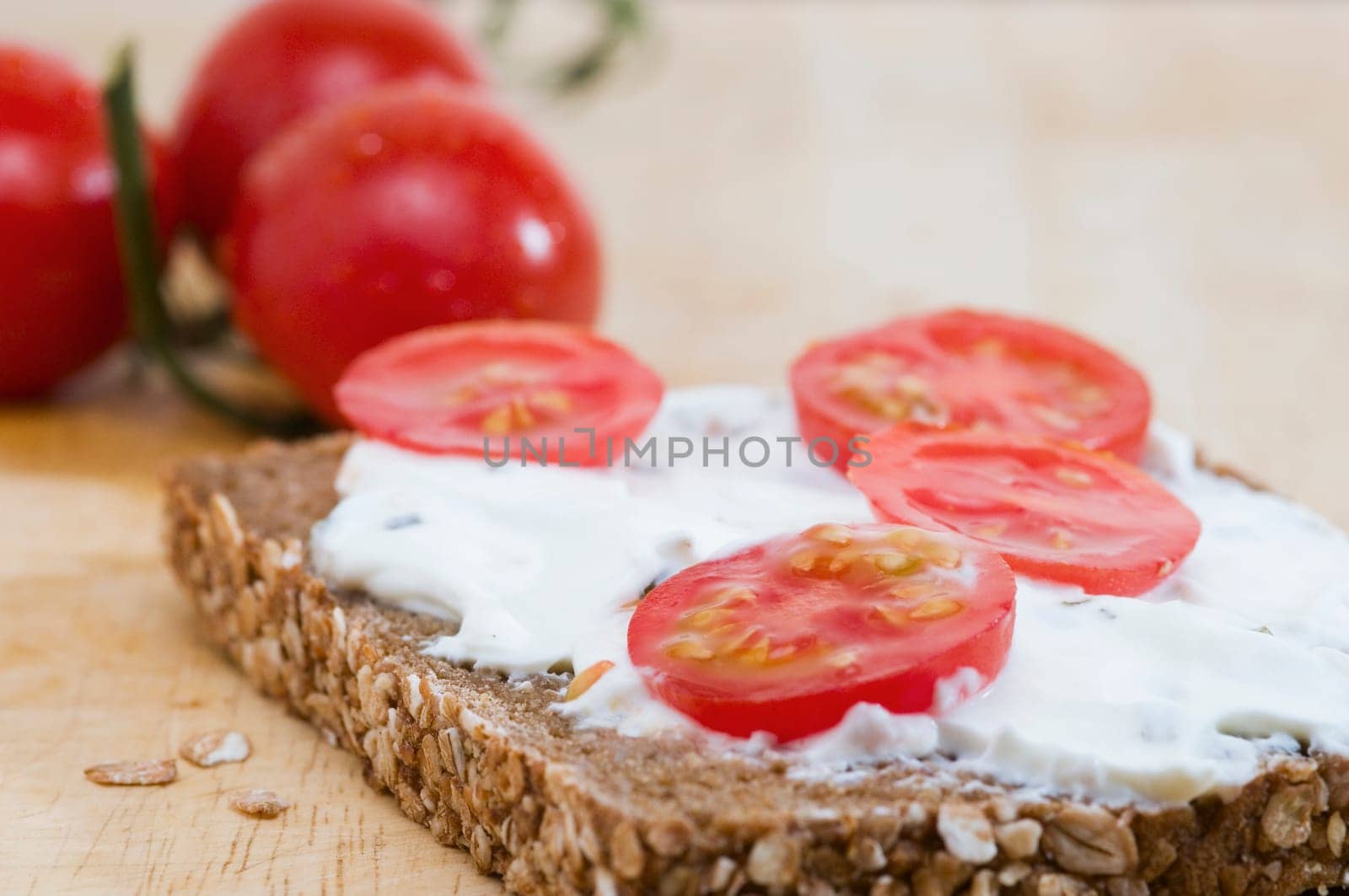 Close-up of a slice of bread coated with white cheese and three tomatoes lying behind it on a wooden cutting board  four slices of tomato as an additional bread topping are on top of