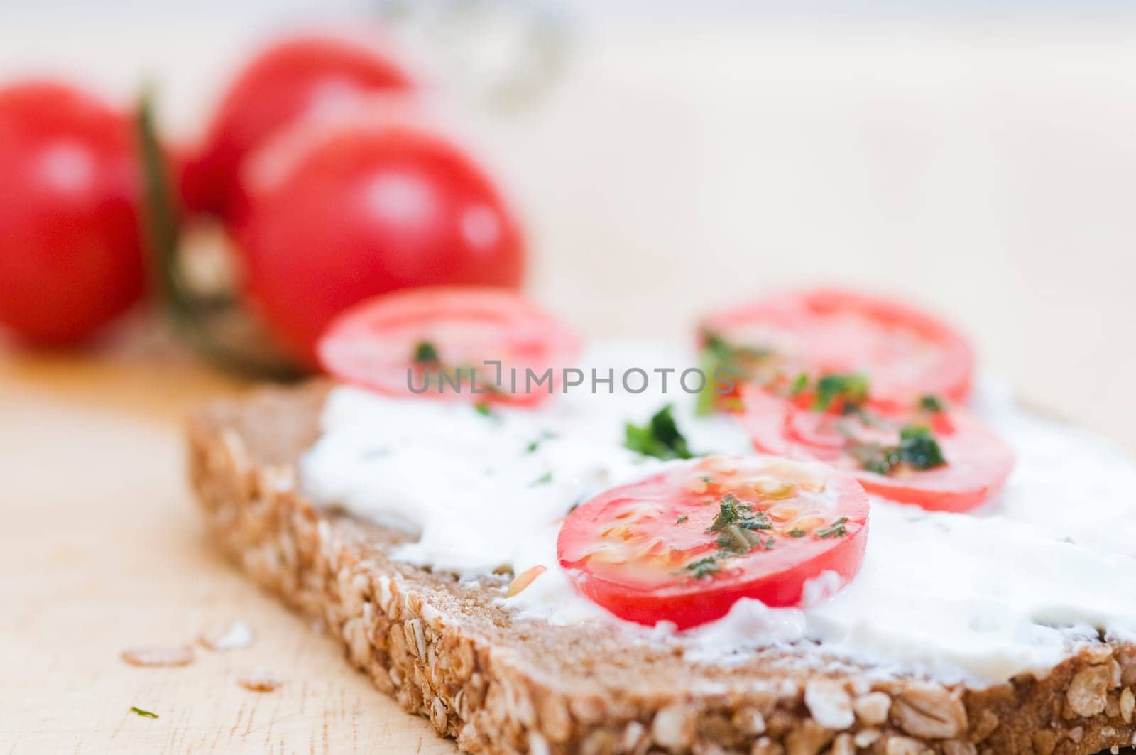 Close-up of a slice of bread coated with white cheese and three tomatoes lying behind it in a fuzzy background on a wooden cutting board  four slices of tomato and slightly chopped herbs as an additional bread topping are on top of