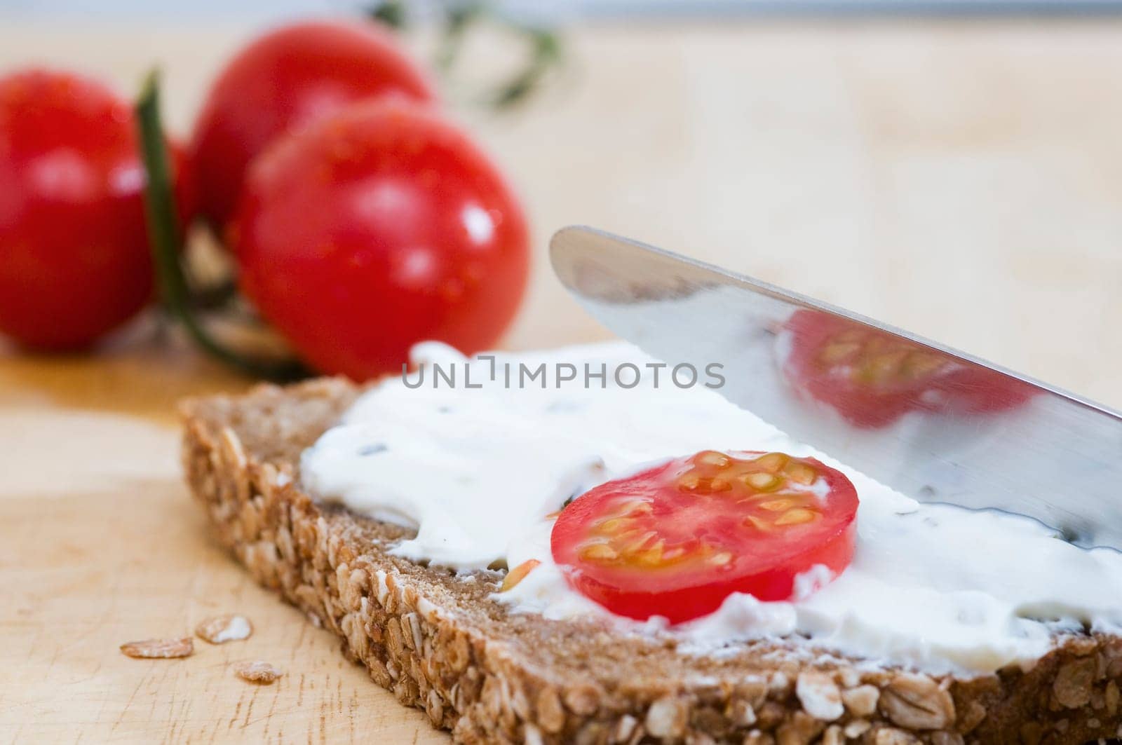 Close-up of a slice of bread coated with a knife of white cheese and two tomatoes lying behind it on a wooden cutting board  a tomato slice as an additional bread topping