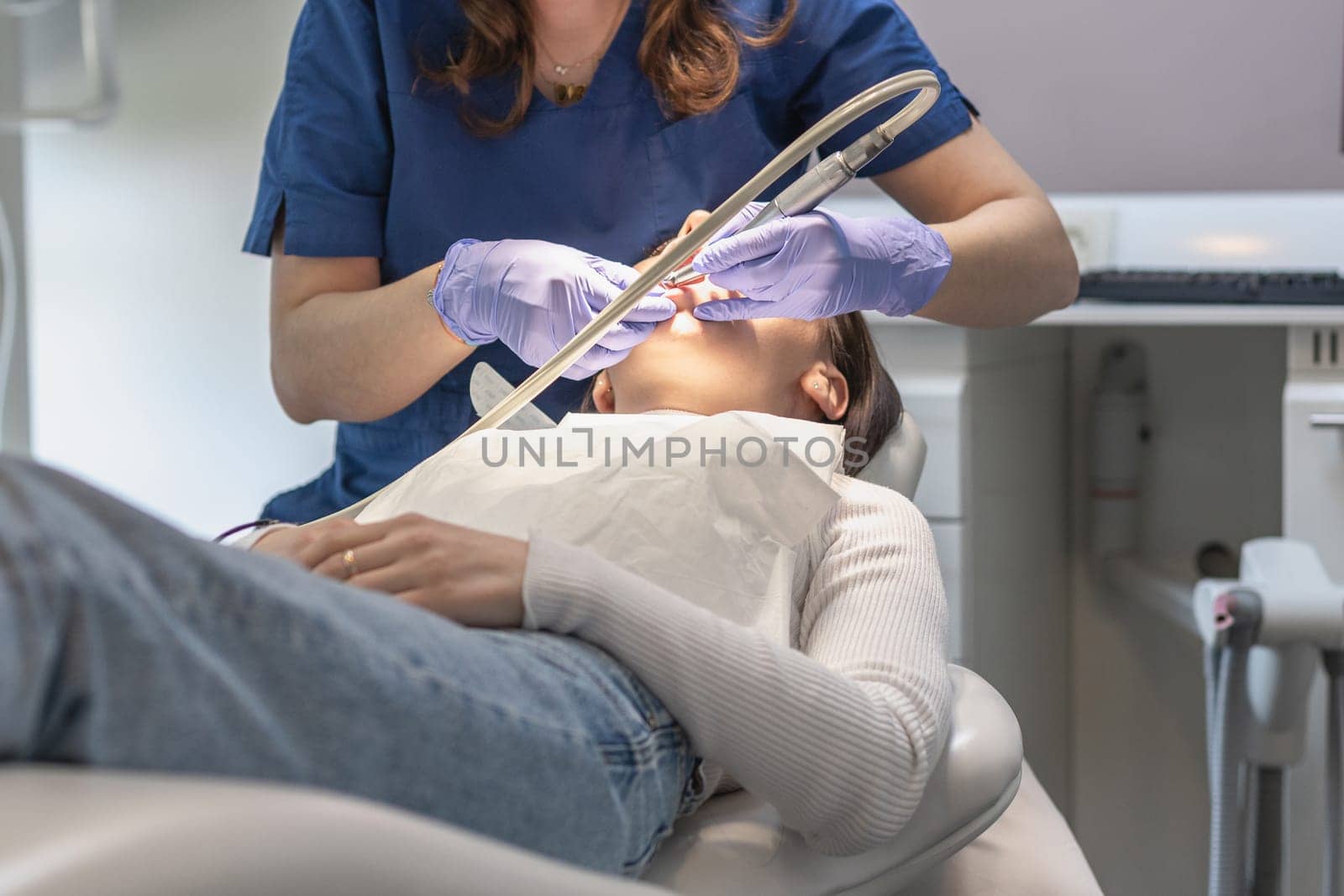A dentist in a blue uniform and lilac gloves performs a deep cleaning of the oral cavity using dental tools of a young brunette patient lying in a chair, close-up view from below. Concept, health and oral hygiene.
