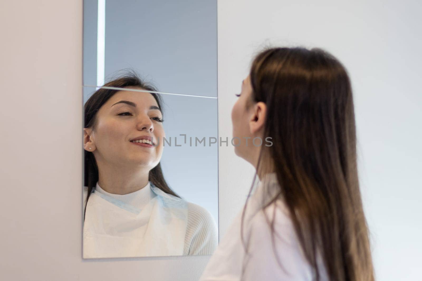 One beautiful young caucasian brunette girl with a smile examines her bleached teeth in the mirror on the wall in the dentist's office in the hospital, close-up side view.