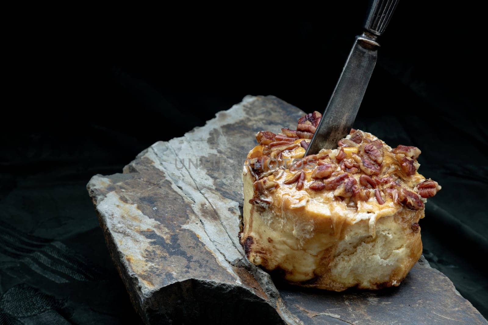 The black metal knife is inserted into the Caramel Pecan topped with caramel frosting and pecans on rock with dark background. Copy space, Selective focus.