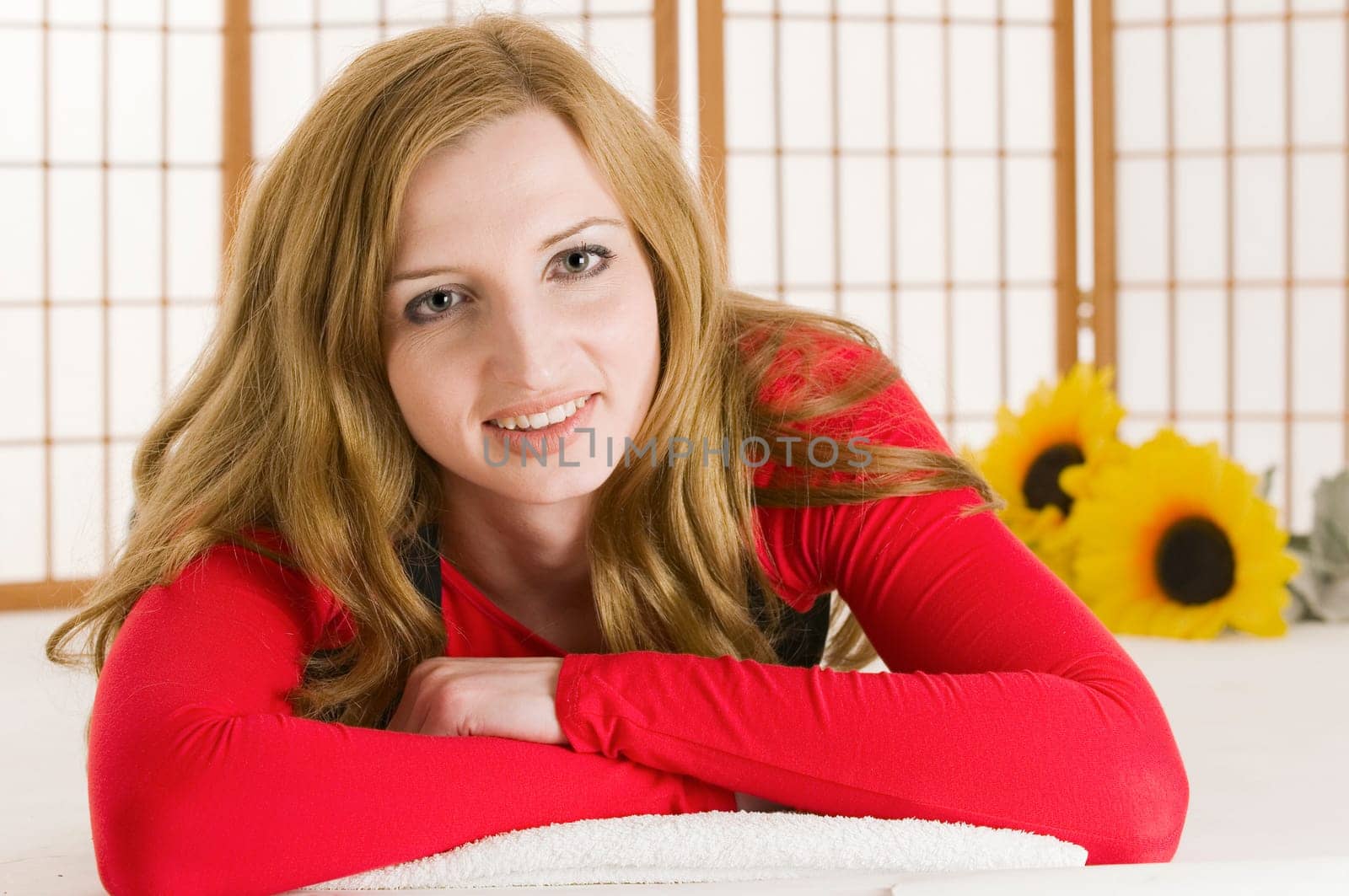 Head-and-shoulder portrait of a young dark blonde woman in a red sweater with her arms crossed on her stomach and looking friendly at the camera