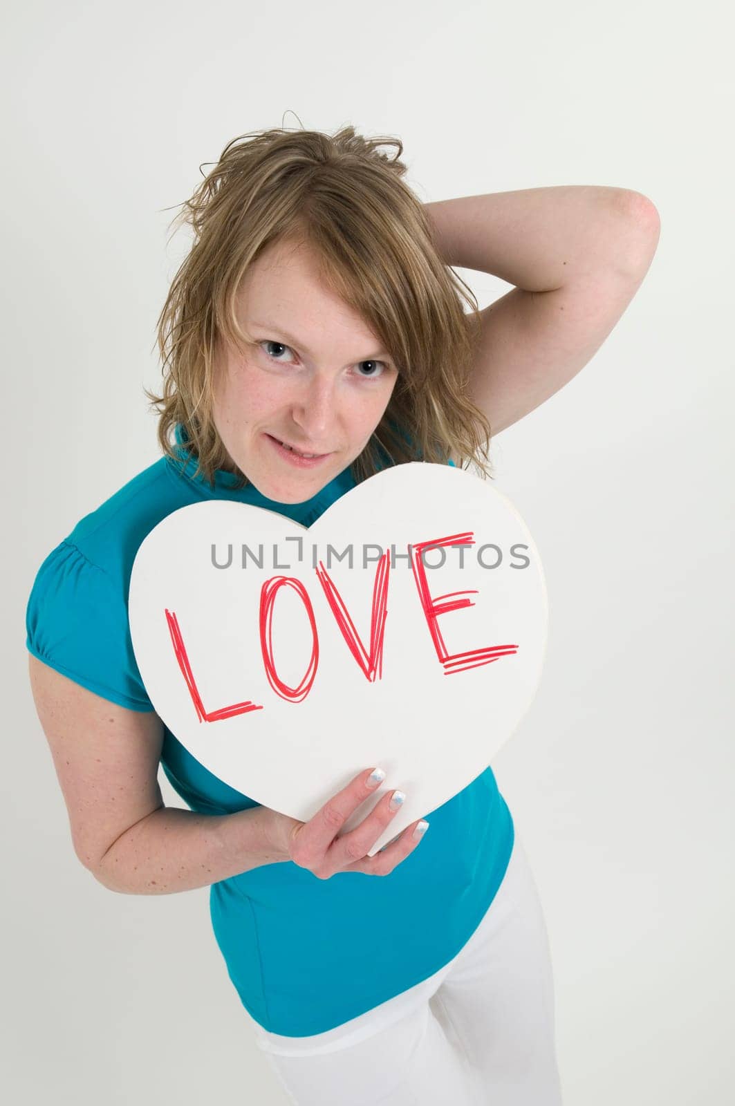 Portrait of a young blonde woman in a light blue T-shirt holding a white heart in front of her with the inscription "LOVE"" and with her left hand reaching into her own hair"