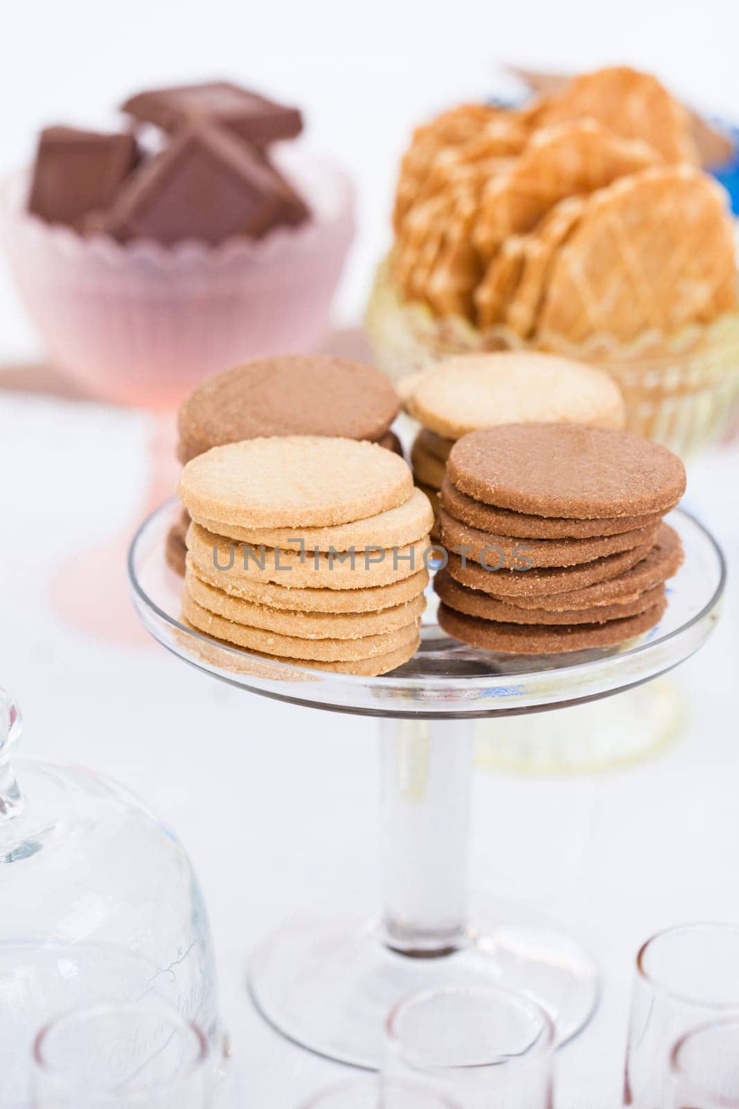 Closeup of four stacks of dry biscuits on glass serving stand with chocolate pieces and waffle cookies in bowls in background
