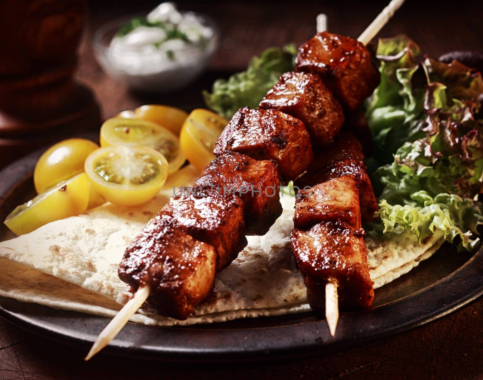 Healthy barbecued lean cubed pork kebabs served with a corn tortilla and fresh lettuce and tomato salad, close up view on a dark background
