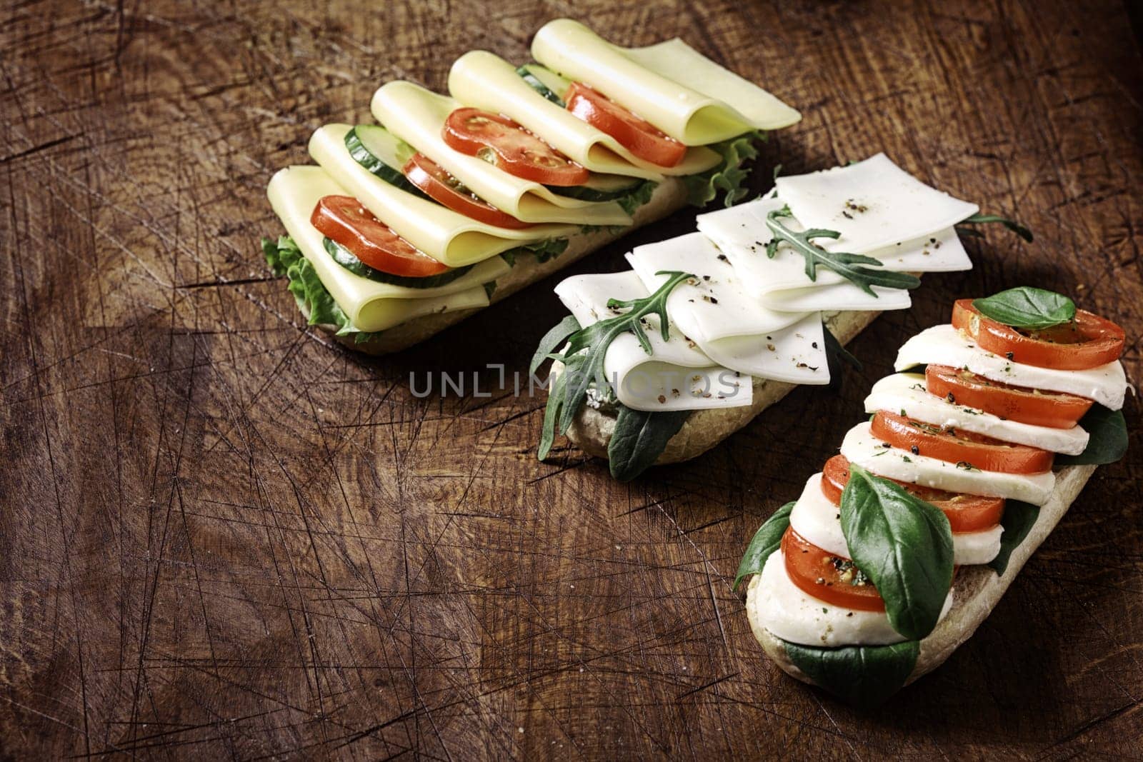Delicious open vegetarian baguettes topped with assorted cheese including gouda and mozzarella with tomato, lettuce, rocket and basil arranged on a rustic wooden table with copyspace, high angle view