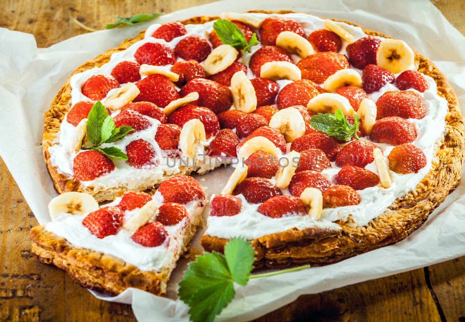 Delicious strawberry and banana pie or tart on a crisp golden crust topped with fresh fruit and cream served with one slice ready to eat, close up high angle
