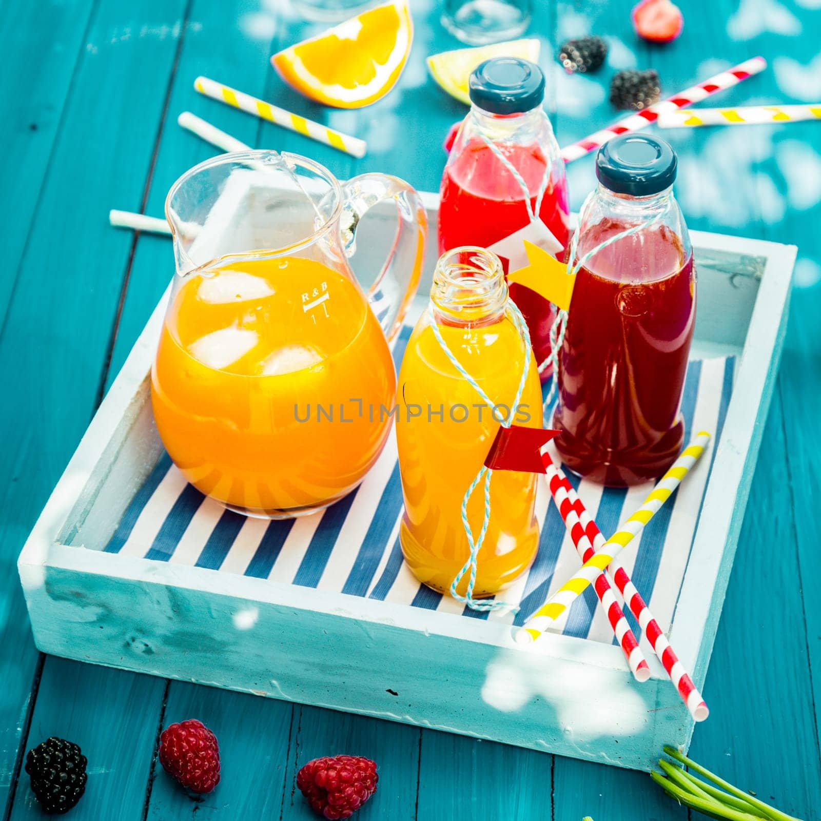 Bottles of freshly squeezed orange and berry juice standing on a wooden tray on a colorful turquoise blue picnic table in dappled summer sun