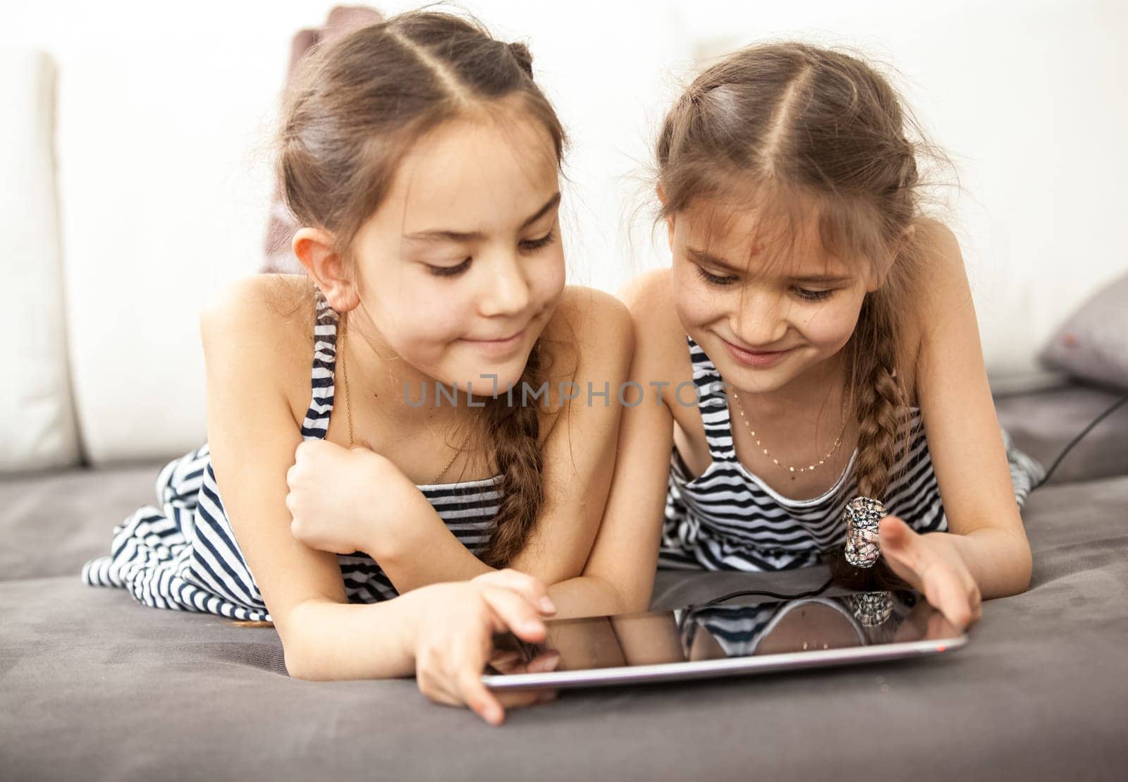 Photo of smiling schoolgirls lying on couch and playing on tablet