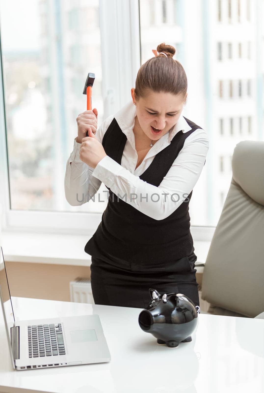 Portrait of angry woman at office hitting piggy bank with hammer