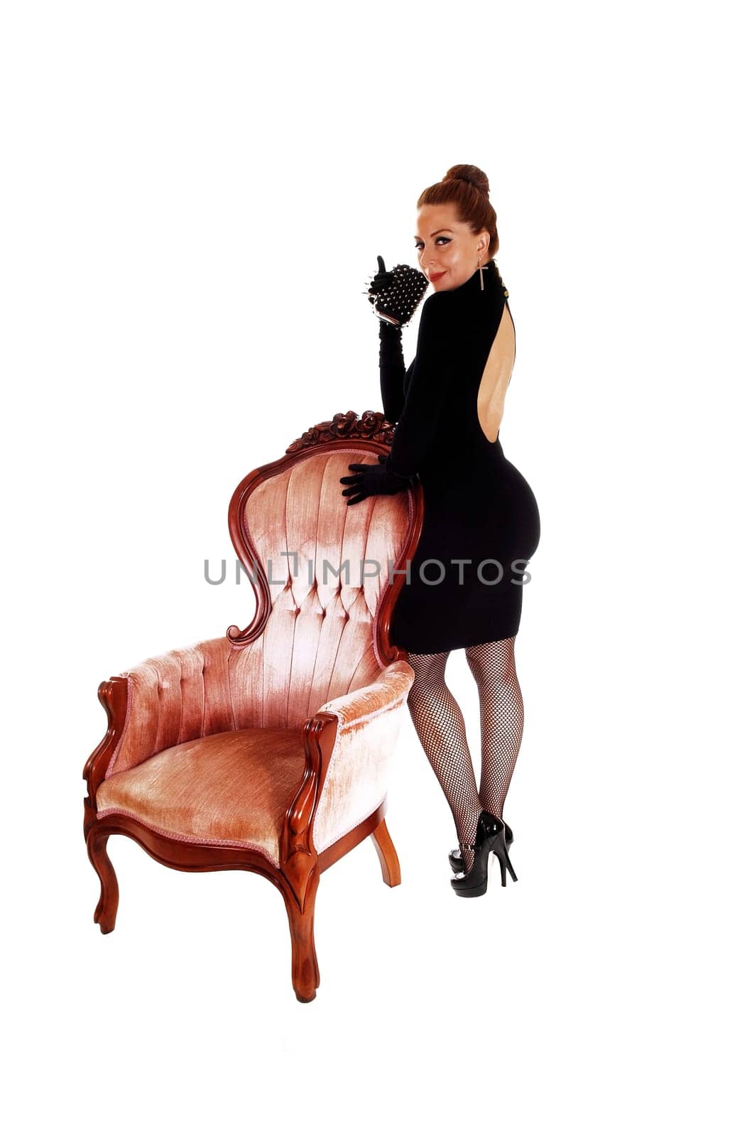 A lovely slim woman in a black dress and high heels standing on a pink.armchair, isolated for white background..