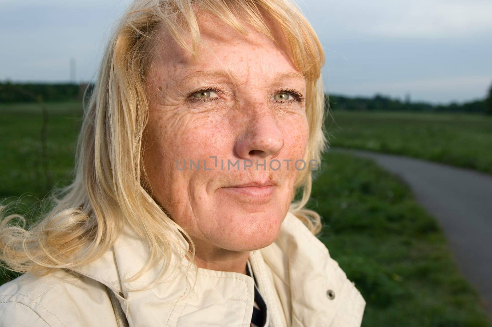 Head-and-shoulder portrait of a blonde mature woman in bright jacket looking at the camera on a green lawn with footpath