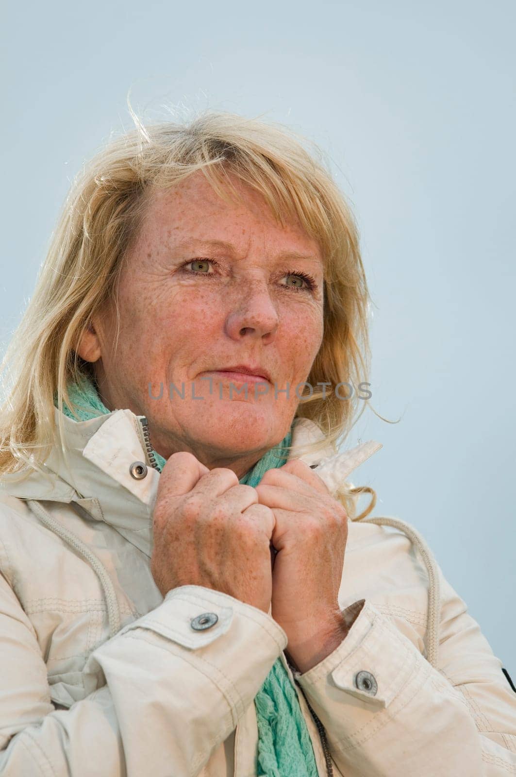 Torso portrait of a blonde mature woman in bright jacket in front of covered sky dreamily looking to the side with her hands at the collar increased view