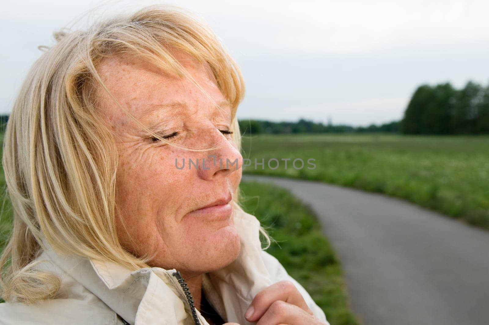 side close-up of a blonde mature woman on a green meadow with her eyes closed deeply inhaling and enjoying the fresh air