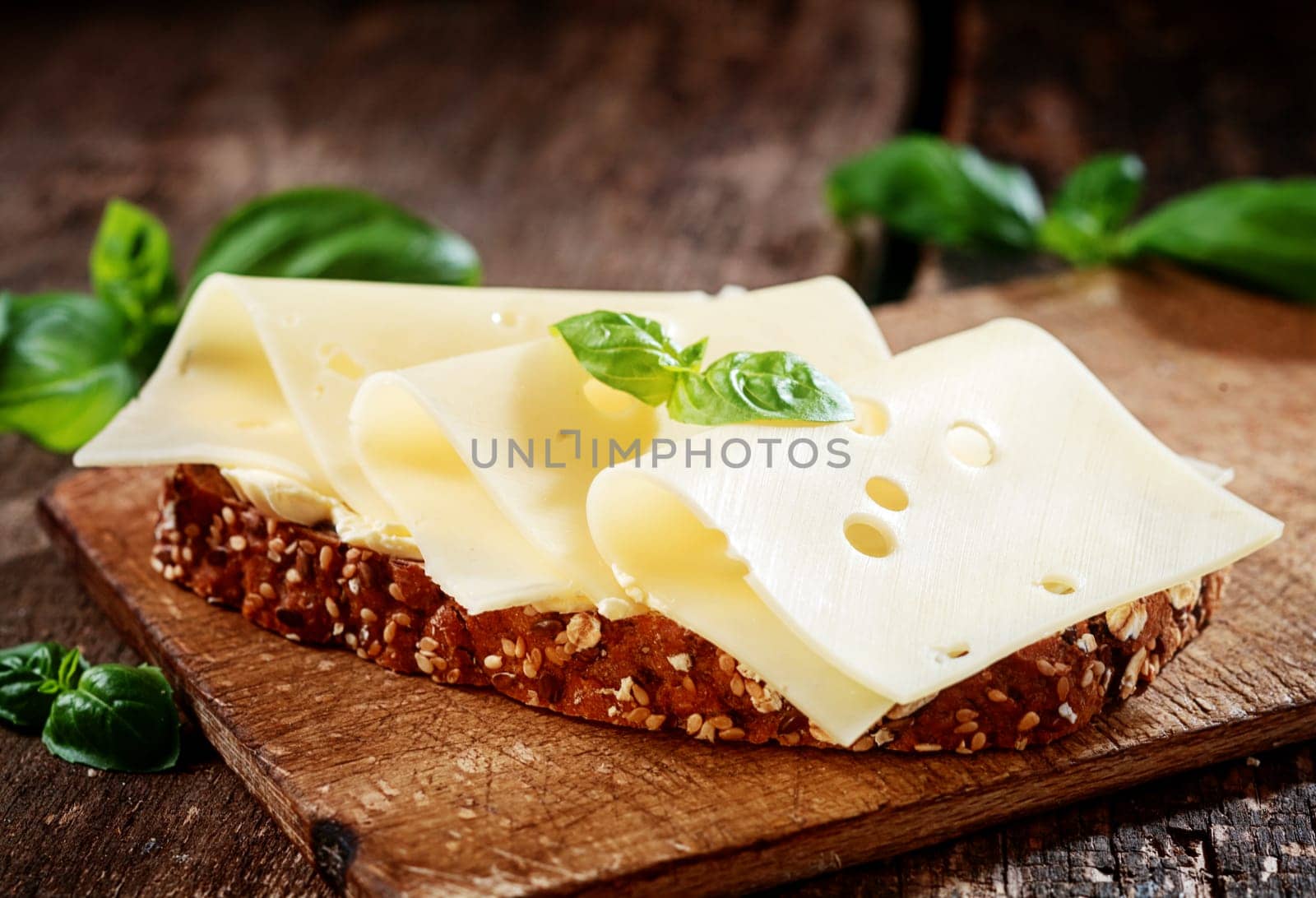 Thinly sliced gouda cheese on wholewheat bread garnished with fresh basil served on a wooden board, close up view