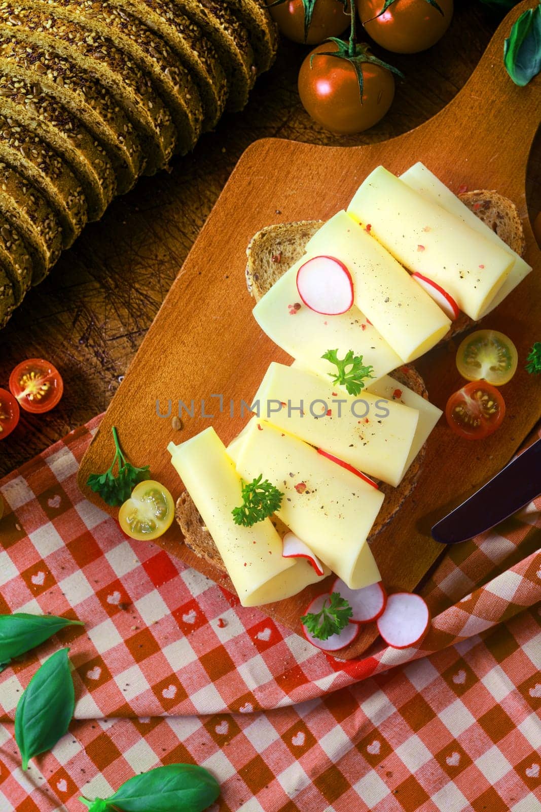 Preparing a delicious gouda sandwich in a country kitchen on a red and white checked cloth with neatly rolled cheese slices on bread with radish and parsley, overhead view