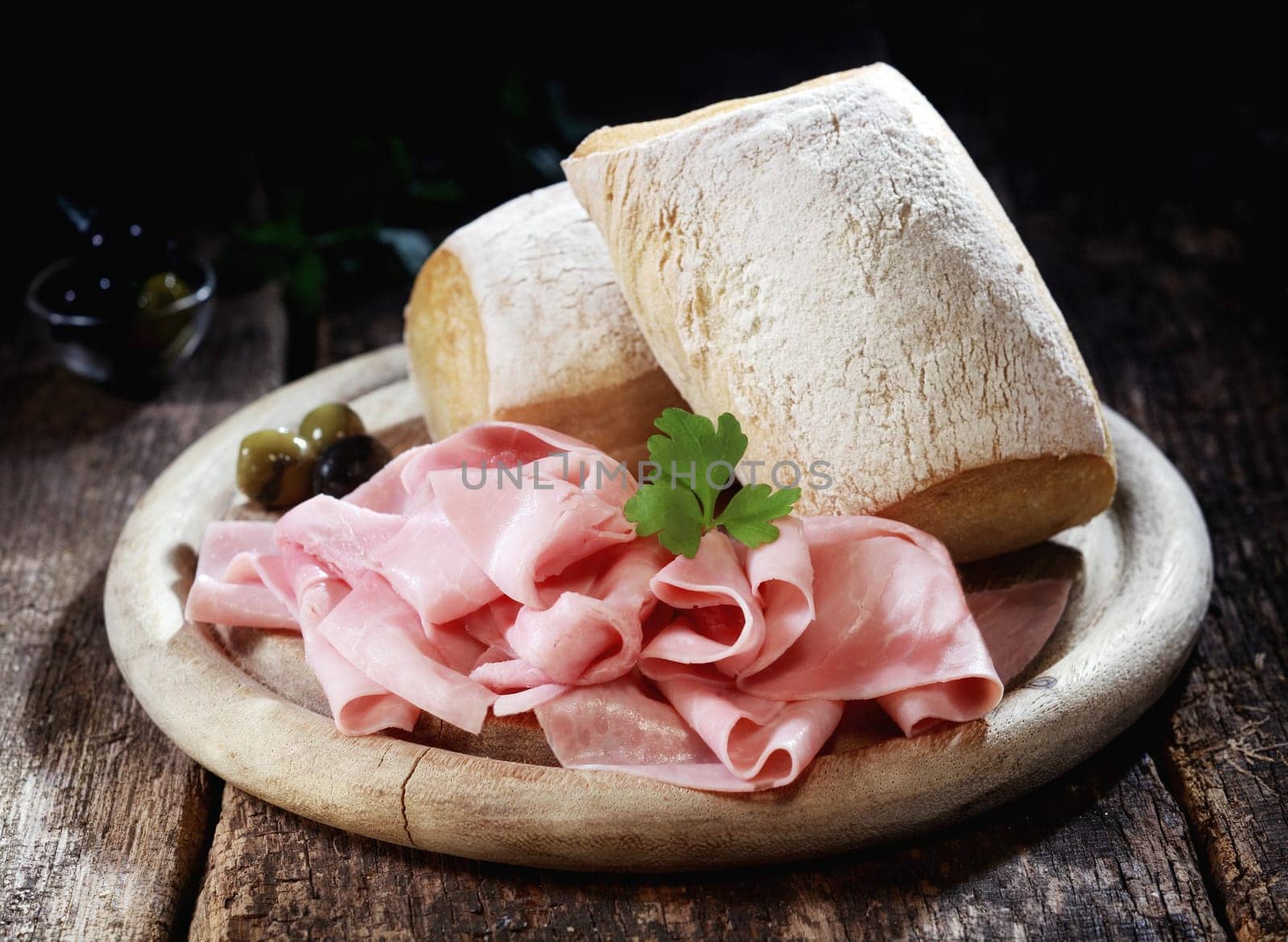 Country lunch with thinly sliced cured Bavarian ham and crusty rolls served on a wooden platter on a rustic wooden table