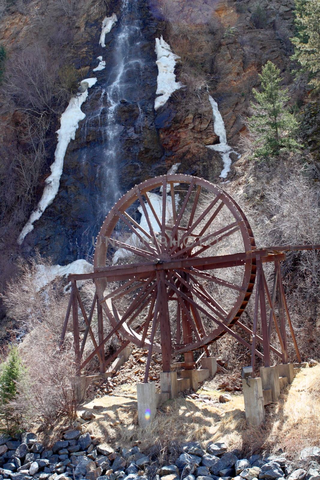 The Charlie Tayler Waterwheel with Bridal Veil waterfall in the background cascading down the mountainside into Clear Creek River on a spring day.