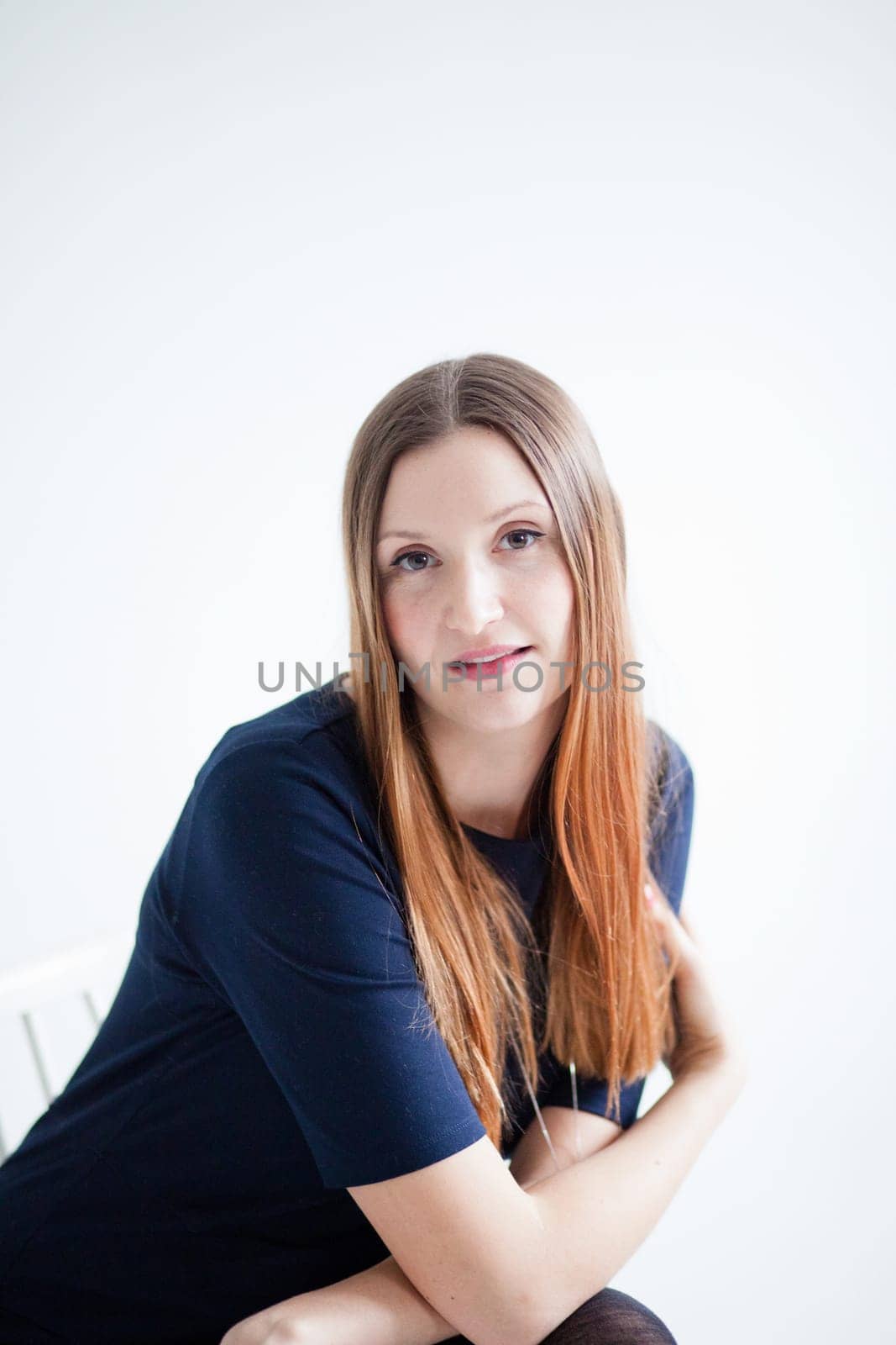 Studio portrait of thoughtful looking attractive woman wearing blue blouse and sitting in chair with arms folded