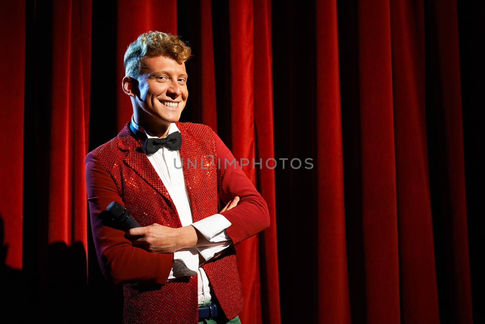 Arts and entertainment in theatre with funny man working as anchorman, standing with arms crossed against red curtains with microphone