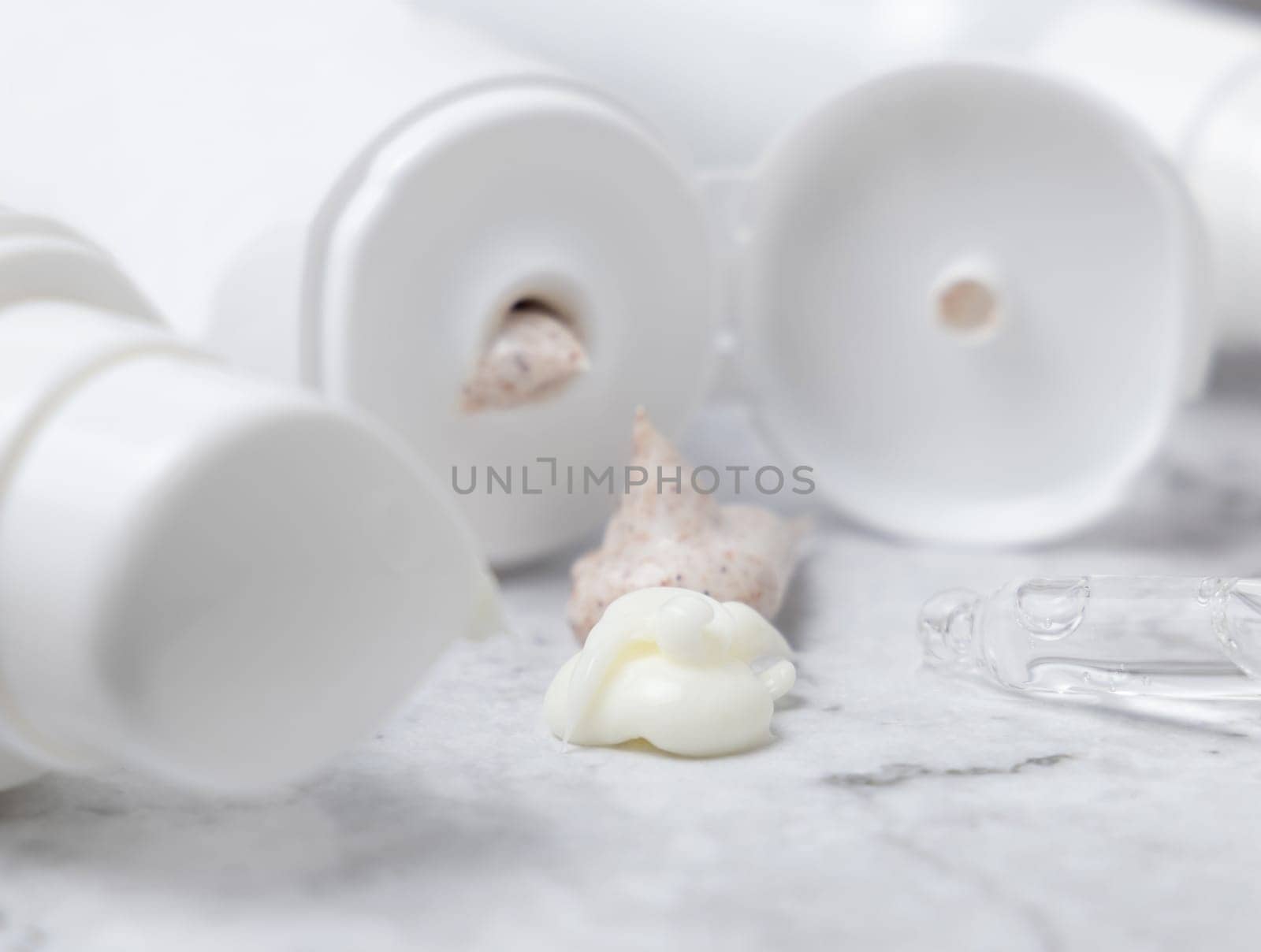 White tube and bottle with open cap and beige cream and scrub squeezed on white marble table, against cosmetic bottles, close up. Natural product for daily face care, spa procedures