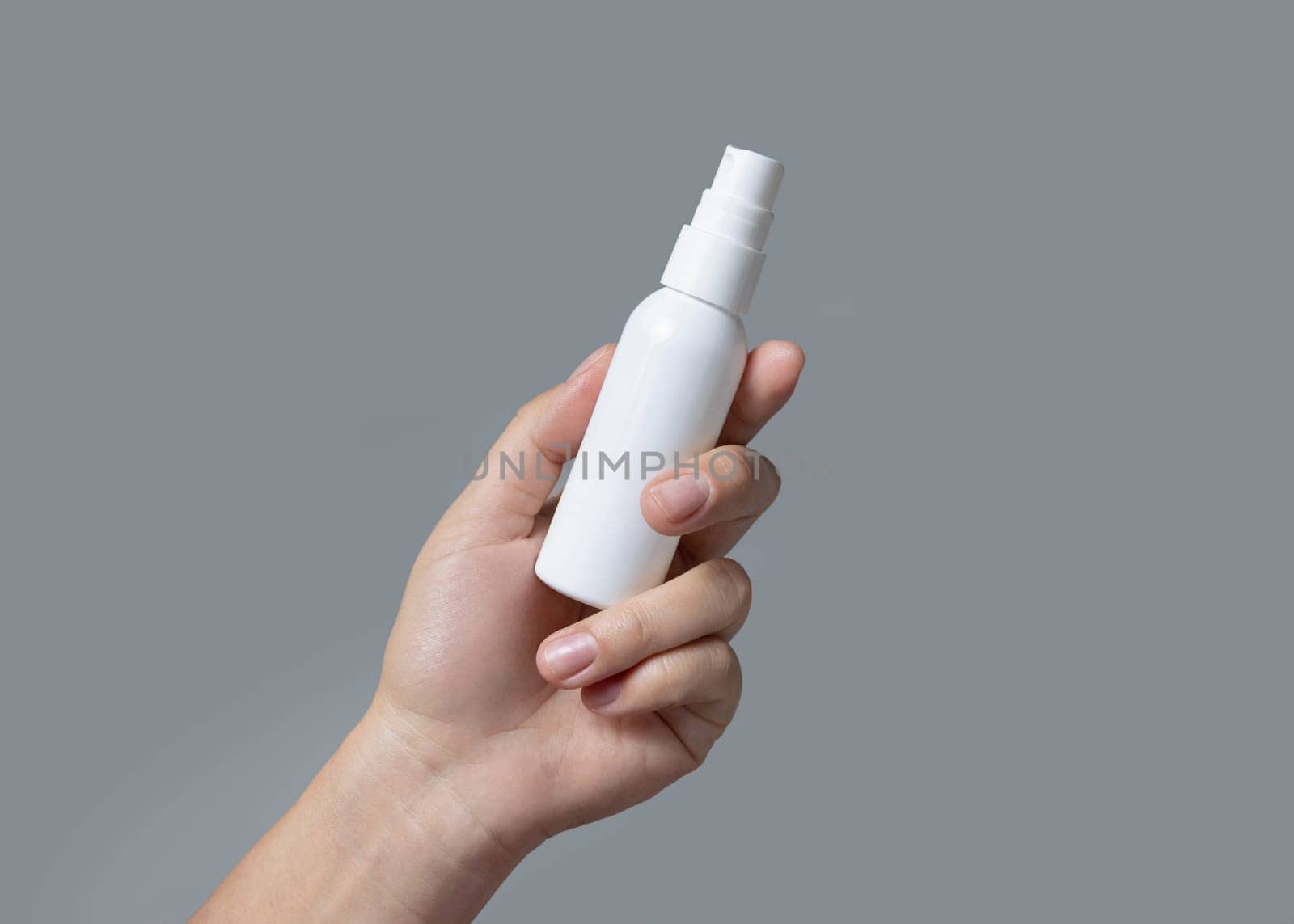 Female hand holding white cosmetic spray bottle against grey close up, package mockup, copy space. Skincare beauty product - lotion or cream