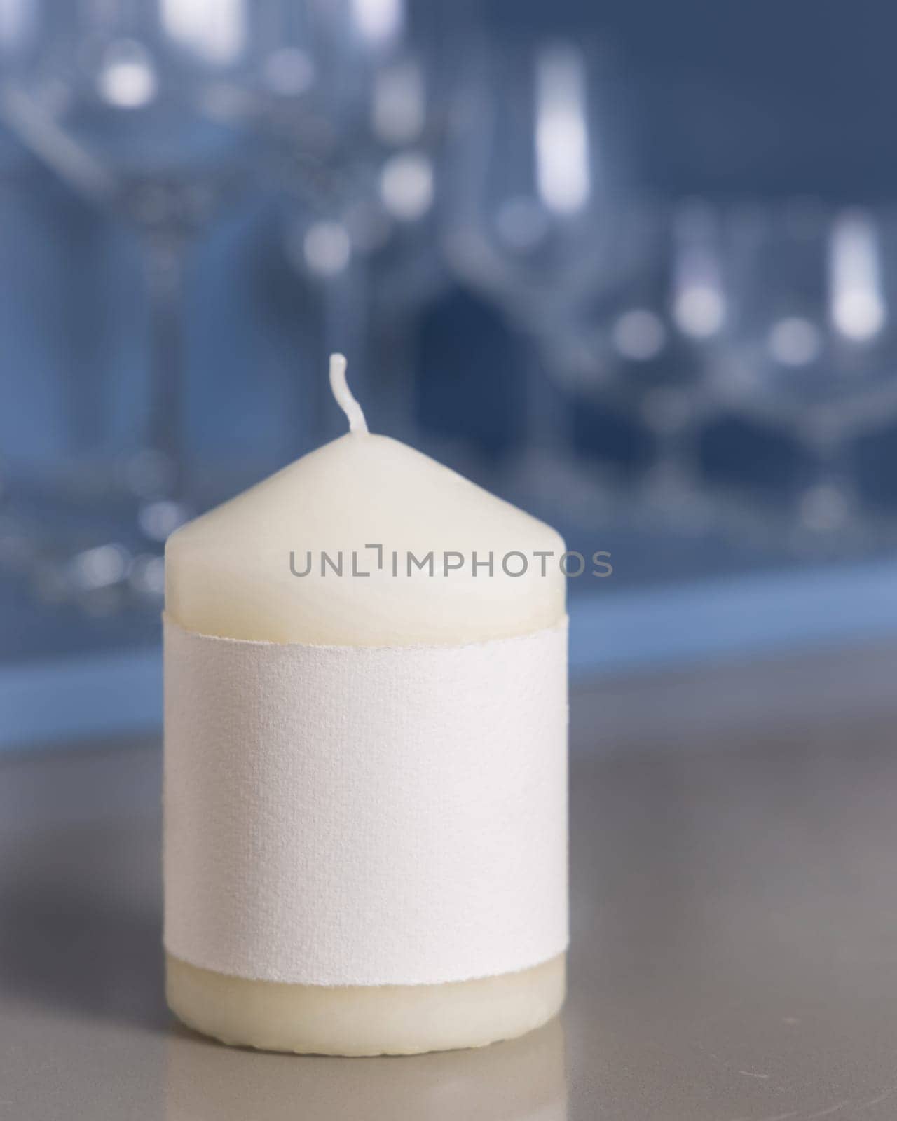 Cream pillar candle with a label on grey kitchen table over blue holder with wine glasses closeup, mockup. Template for advertising and design with copy space