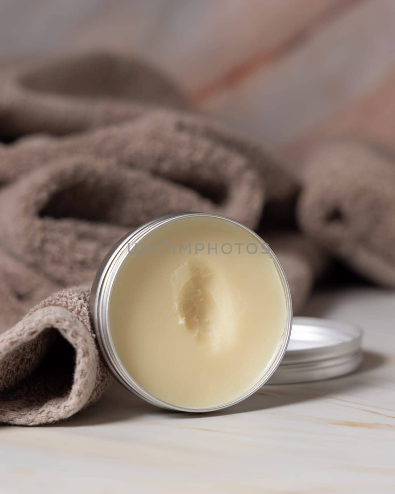 Opened body butter jar near light brown bath towel in bathroom, close up. Natural herbal cosmetic product for body care. Daily beauty routine or spa procedures